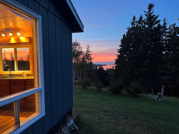 Een huis met een zichtbare keuken, een raam met uitzicht op een gazon, en een beeld van bomen en een zonsondergang.