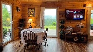 A wooden interior with a dining table, chairs, a TV, and a view of greenery outside. - Margaree-a-ville Cottage (Margaree Harbour)