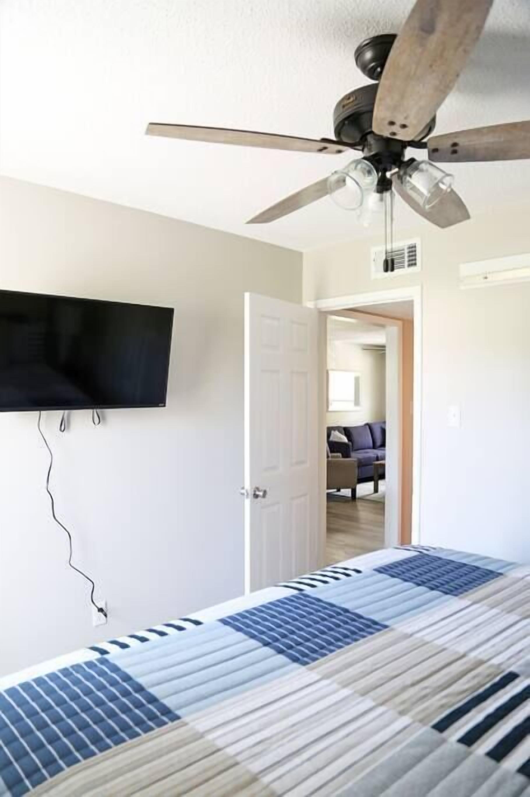 Un dormitorio con una cama, ventilador de techo, televisor montado y una puerta que da a otra habitación.
