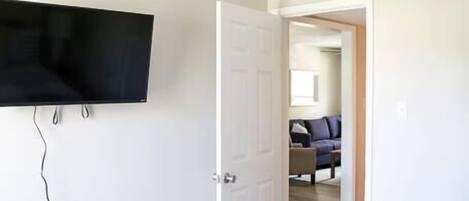 Un dormitorio con una cama, ventilador de techo, televisor montado y una puerta que da a otra habitación.