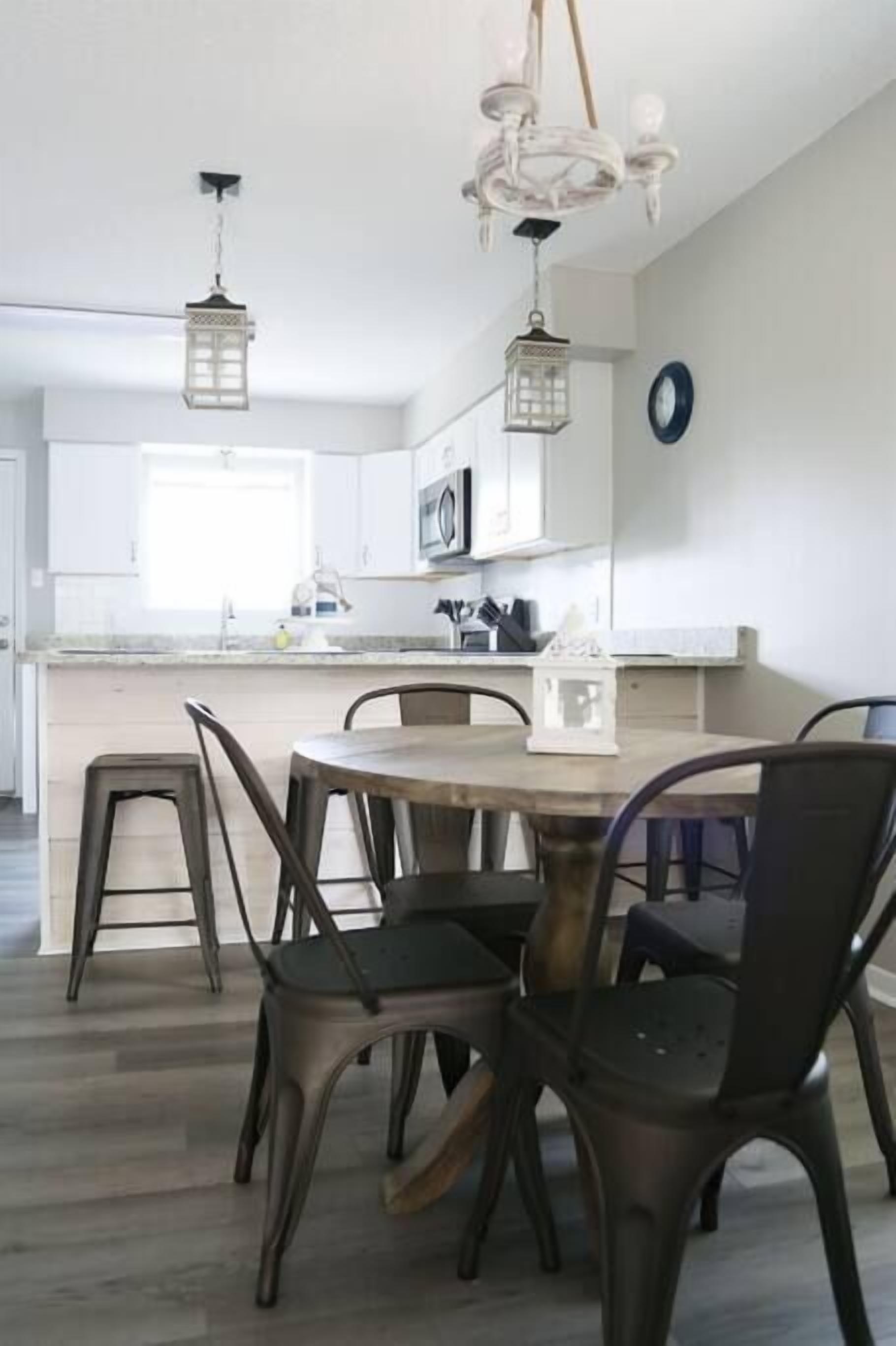 A modern kitchen with white cabinets, a wooden dining table, and metal chairs. There are two hanging lantern-style lights above the table.