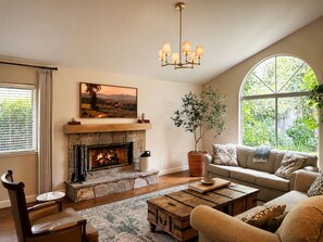 A living room with a fireplace, a sofa, a coffee table, and a large window with a view of greenery. - Spacious Oasis Hot Tub Backyard Garden 3BR/2BA (Sonoma)