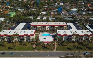 Un hotel con piscina central, rodeado de palmeras y un estacionamiento.