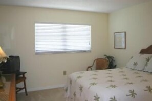 Un dormitorio con cama, escritorio, silla, ventana con persianas y un cuadro en la pared.
