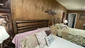 A bedroom with a wooden headboard, a bed with floral bedding, a bedside table with a lamp, and a decorative wall piece. - Hilda’s Garage Bandera, TX (Bandera)