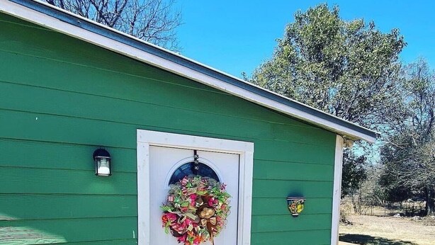 A green shed with a white door, a hanging wreath, and a small light fixture above the door. - Hilda’s Garage Bandera, TX (Bandera)