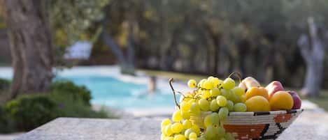 A bowl of fruit on a table with a pool and trees in the background.