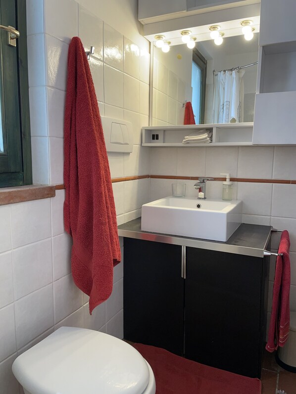 Casa de banho com sanita branca, lavatório branco e espelho sobre o lavatório. Há uma toalha vermelha pendurada na parede e outra no chão.