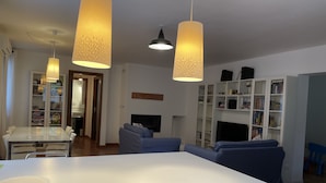 Ein modernes Wohnzimmer mit einem weißen Esstisch, blauen Sofas und einem Fernseher. Zwei Pendelleuchten und ein Bücherregal voller Bücher sind ebenfalls vorhanden.
