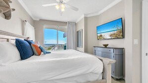 Un dormitorio con una cama grande, ventilador de techo, vista a la playa, cómoda gris y un cuadro de la playa.