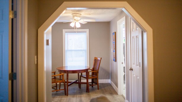 A dining area with a round table and four chairs, a ceiling fan, and a window with blinds.