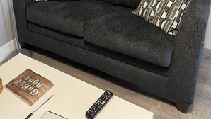 A dark grey sofa with two patterned pillows, a remote control, a book titled 'GUEST BOOK', and a pen on a white table.