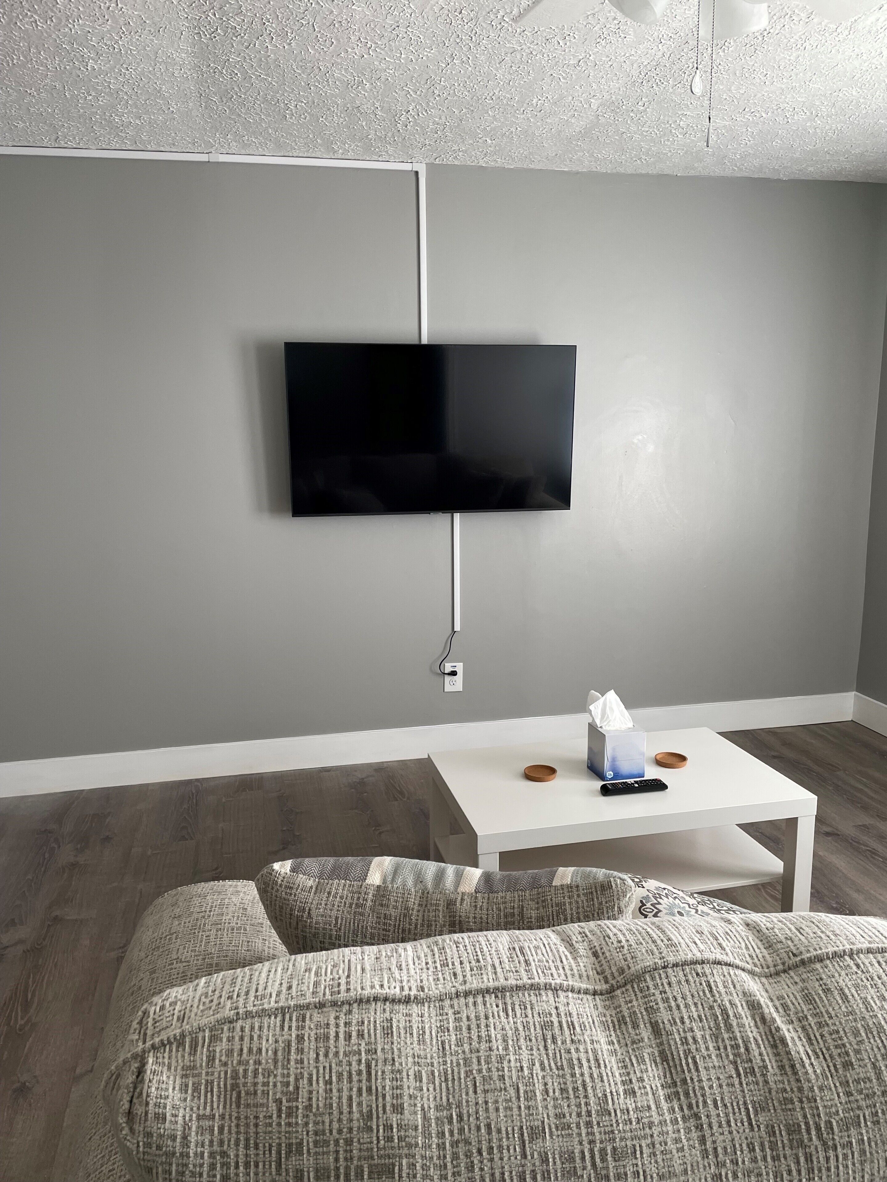 Un salon moderne avec un téléviseur à écran plat fixé au mur, une table basse blanche avec une télécommande et des mouchoirs, et un canapé gris agrémenté de coussins à motifs.