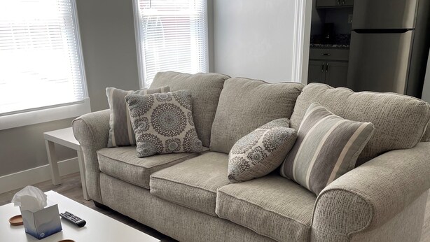 A living room with a beige sofa, a white coffee table, and a kitchen in the background.