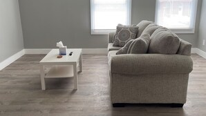 A spacious living room with a grey couch, a small white table, and two windows with blinds.