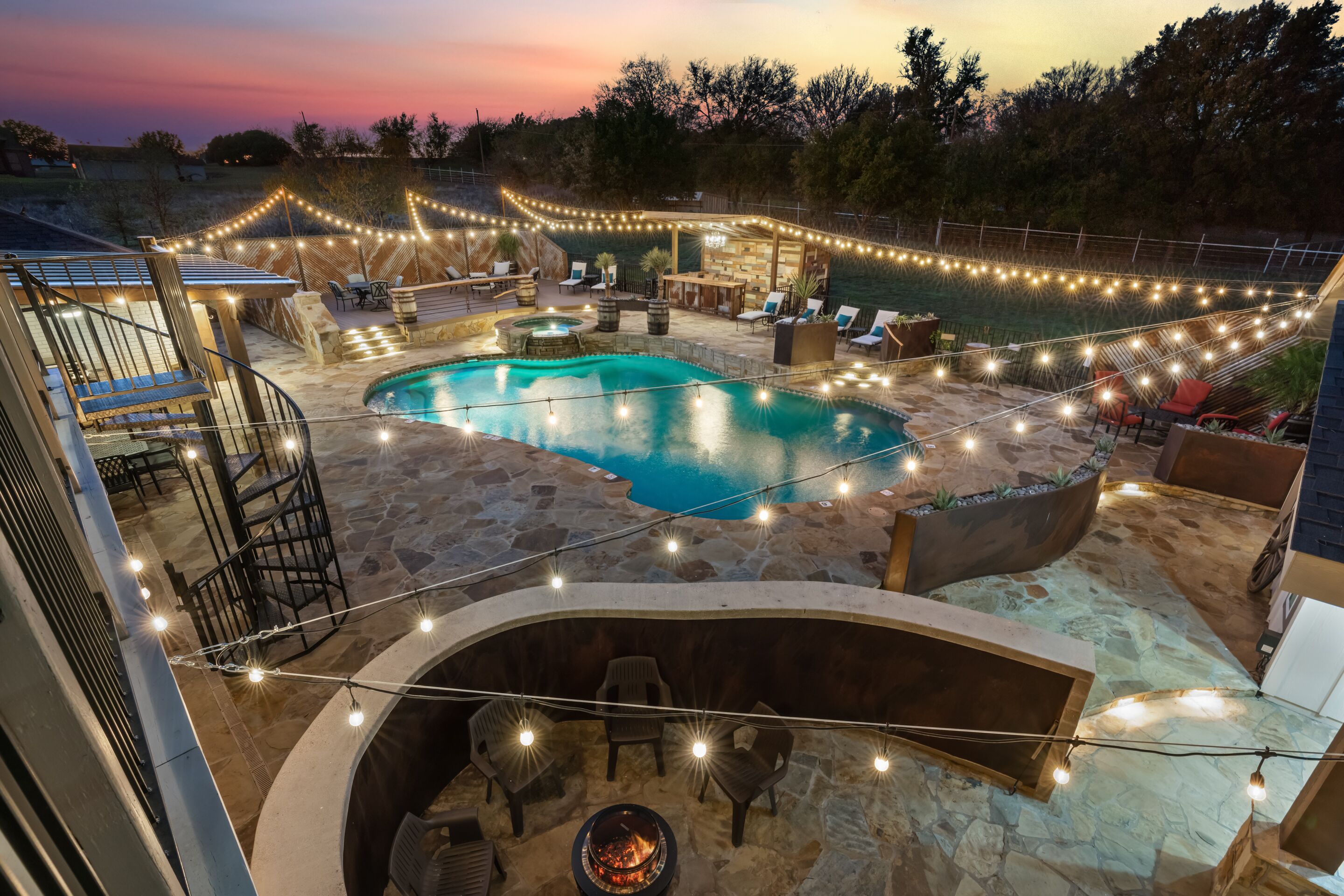 A backyard with a swimming pool, stone patio, outdoor seating, and string lights.