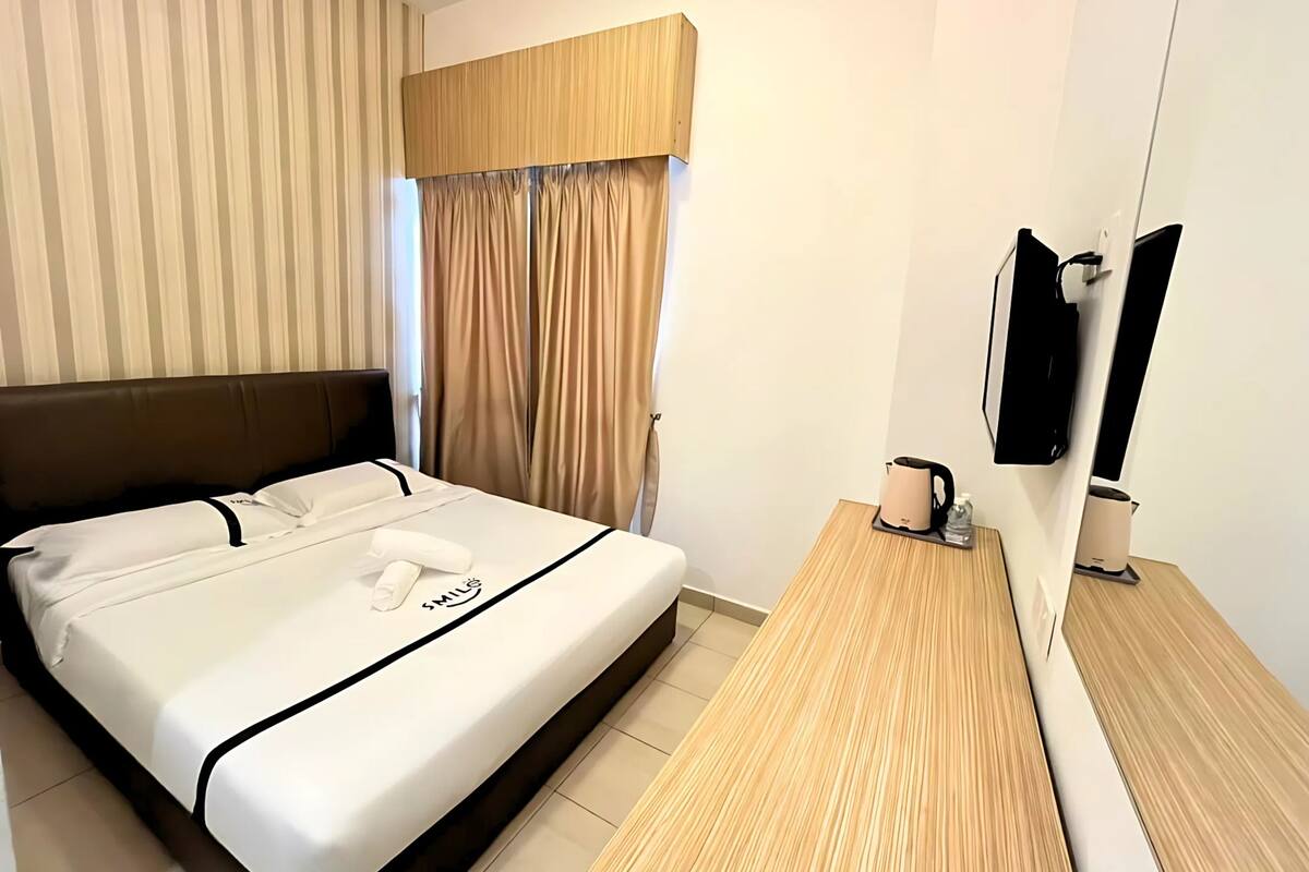 A hotel room with a bed, a wooden nightstand, a kettle, a bottle of water, and a television.