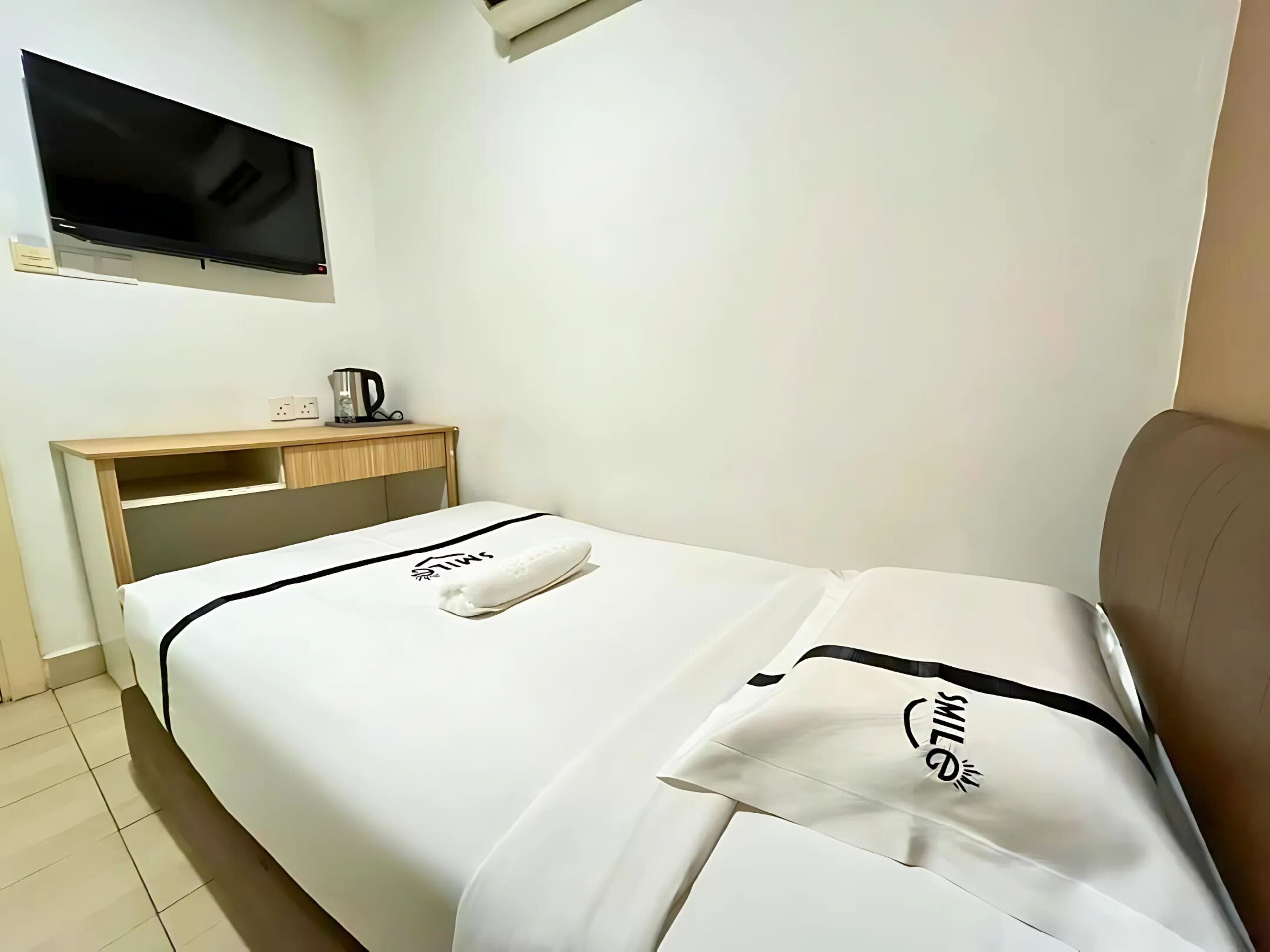 Una habitación de hotel con una cama, un escritorio, un televisor instalado en la pared y una tetera en el escritorio.