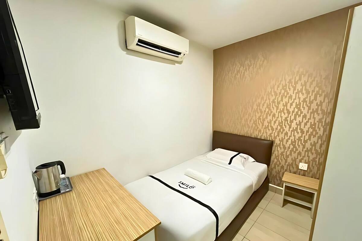 A hotel room with a bed, a small table with a kettle, an air conditioner, and a wall-mounted television.