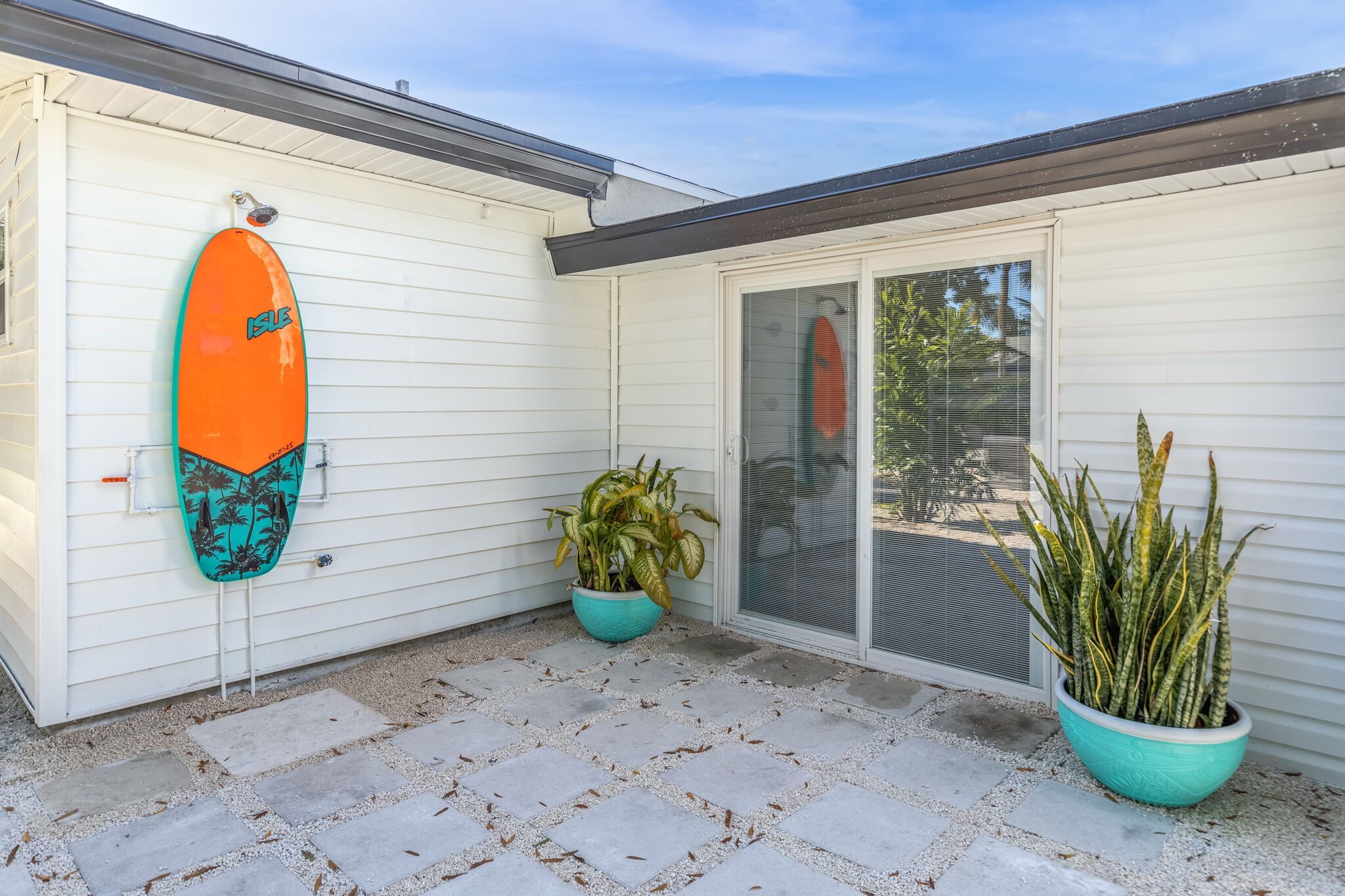 A house with a sliding glass door, a surfboard leaning against the wall, and two potted plants.