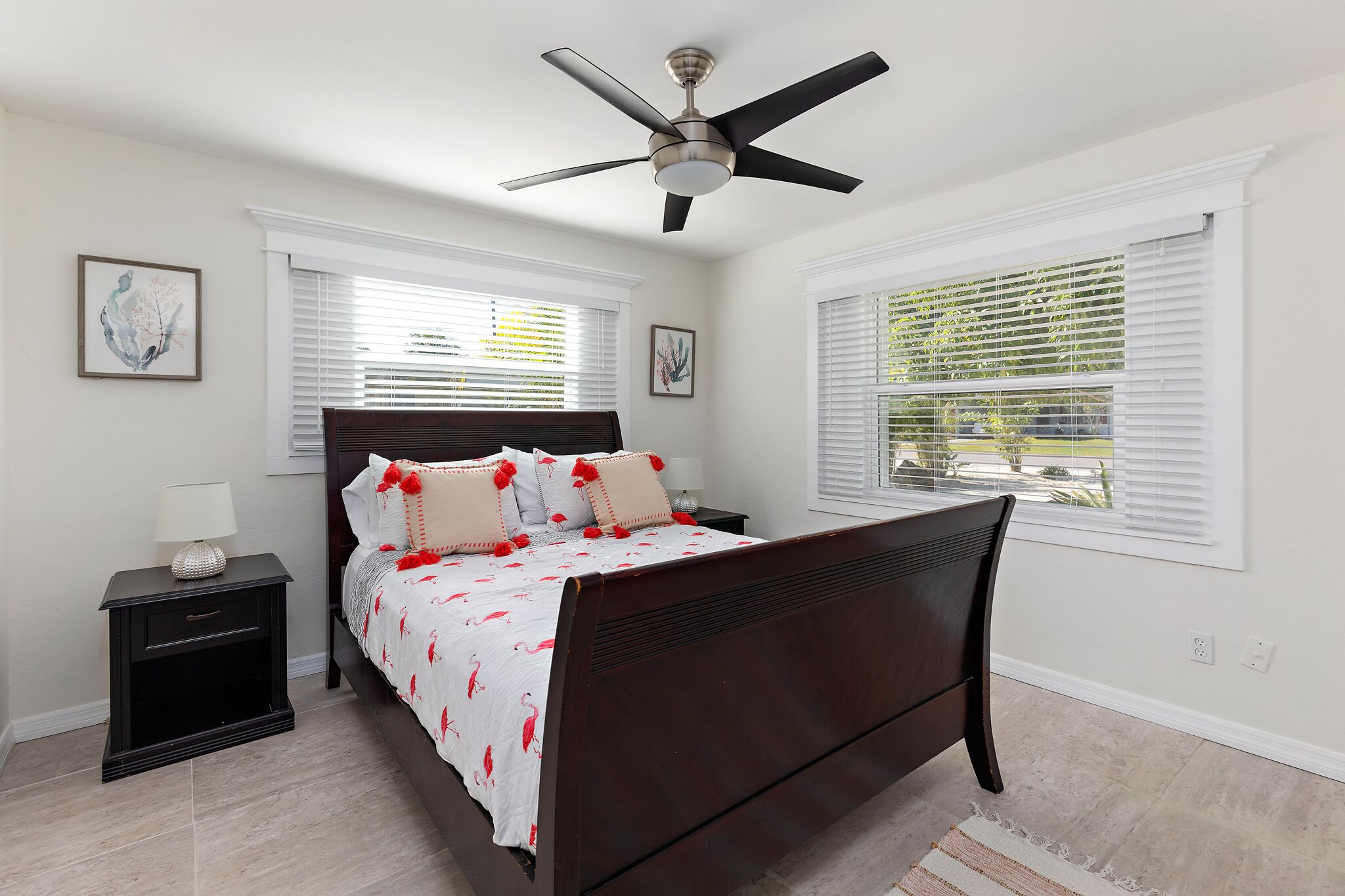A bedroom with a dark wooden bed, white walls, and a ceiling fan. There are two windows with blinds, a nightstand with a lamp, and artwork on the walls.