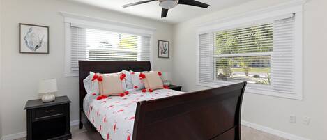 A bedroom with a dark wooden bed, white walls, and a ceiling fan. There are two windows with blinds, a nightstand with a lamp, and artwork on the walls.