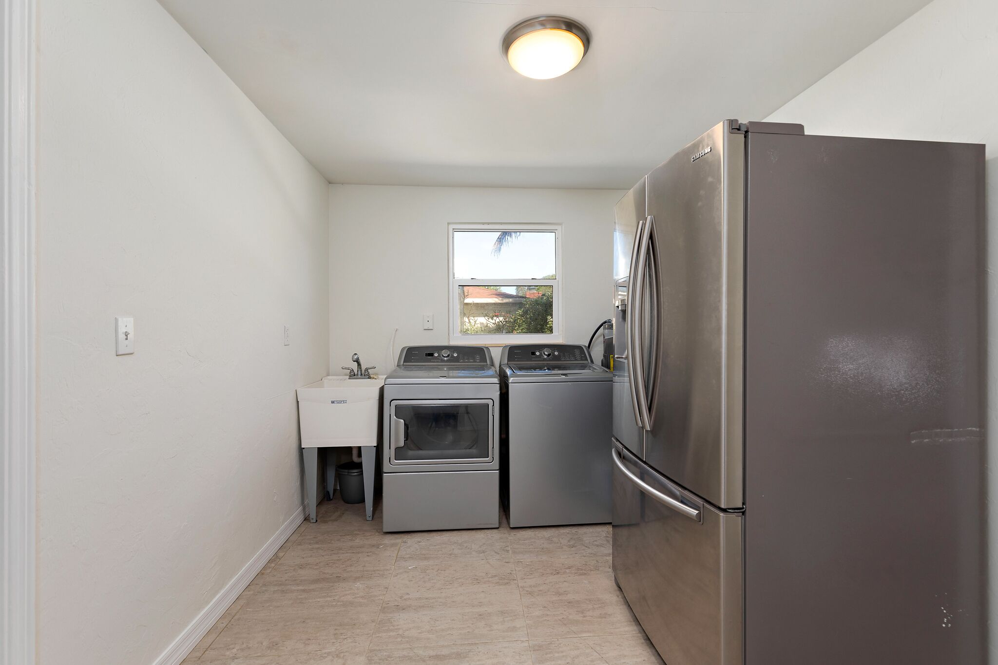 A compact kitchen with a stainless steel refrigerator, two washing machines, a sink, and a window.