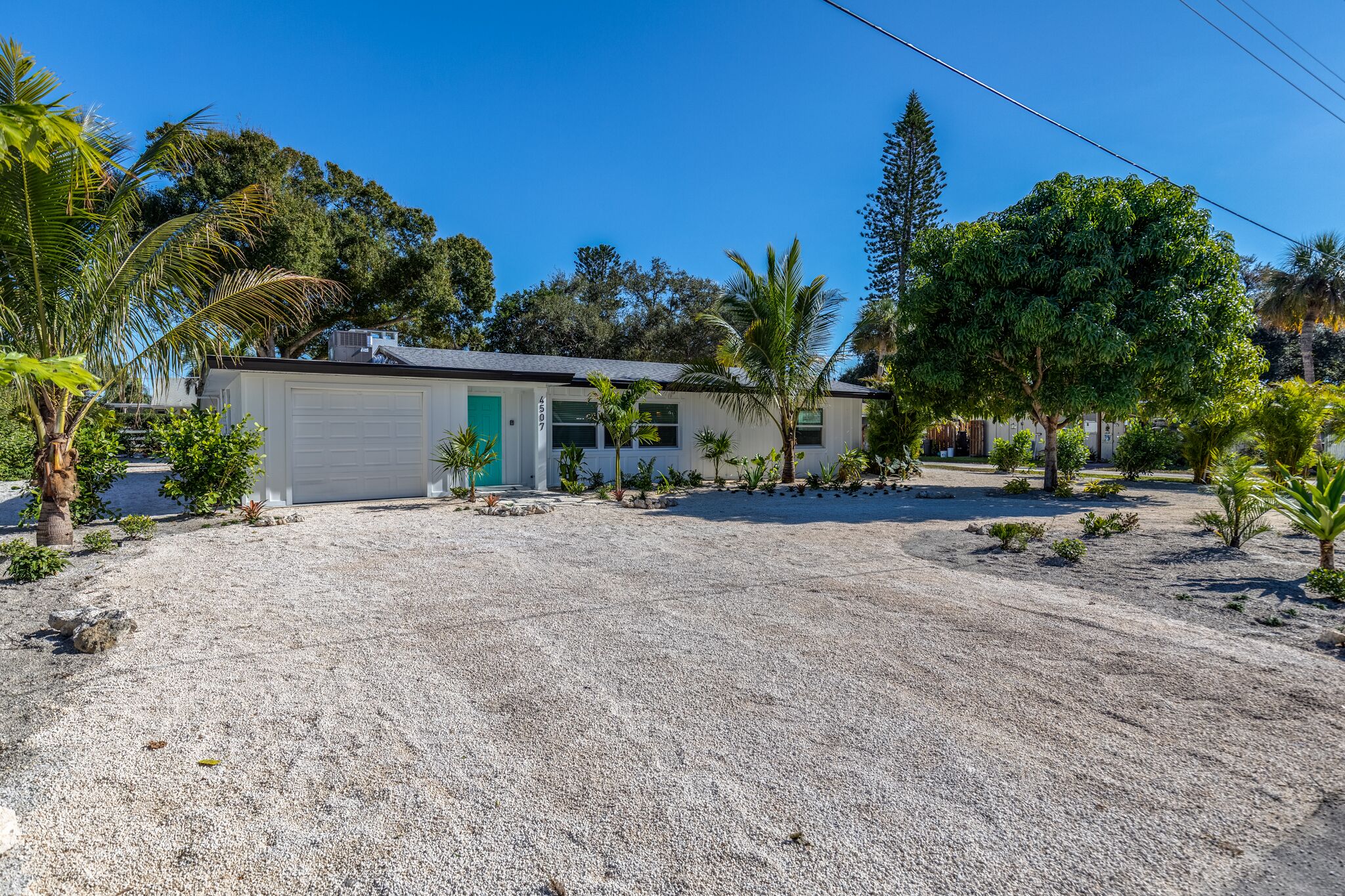 A single-story house with a white garage door, surrounded by a gravel driveway and various small plants and trees.