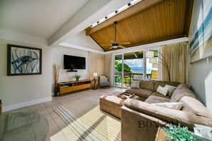A spacious living room with a large sofa, a flat-screen TV, a wooden ceiling, and a balcony with a view of the ocean.