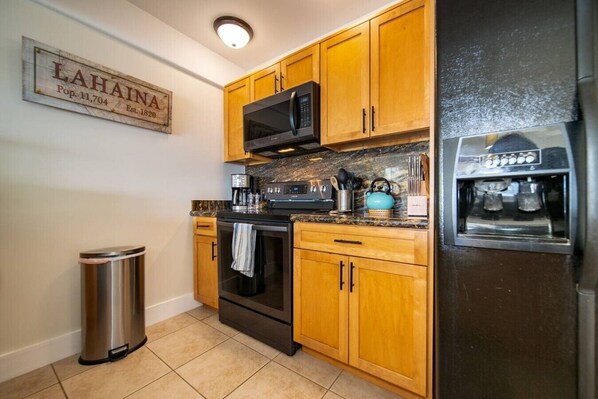 A kitchen with wooden cabinets, a stainless steel trash bin, a black oven, a microwave, and a refrigerator.