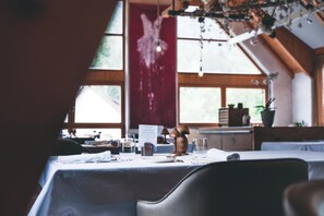 A dining area with tables set for two, a menu stand, and a view of the outdoors through large windows. - Logis Hôtel Frankenbourg (Rocamadour)