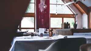 Un área de comedor con mesas dispuestas para dos, un atril para el menú y vistas al exterior a través de amplias ventanas.