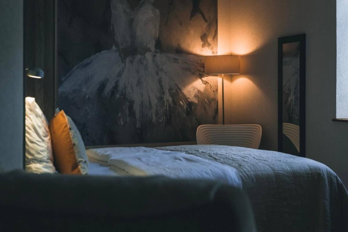 A hotel room with a large abstract painting on the wall, a bedside lamp, a bed with pillows, and a chair.