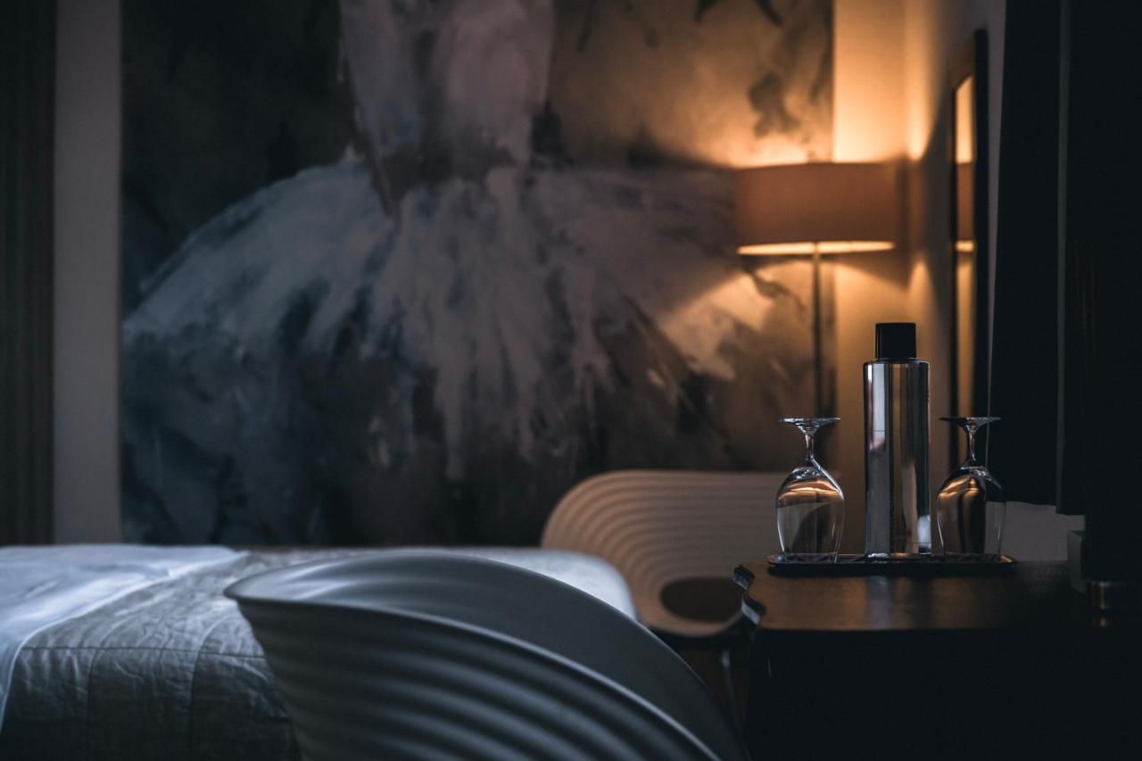 A hotel room with a bed, a bedside table with two wine glasses and a bottle, and a large abstract painting on the wall.