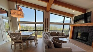 Ein großzügiger Wohnbereich mit einer großen Couch, einem Esstisch, Stühlen, einem Kamin und großen Fenstern mit Blick auf die Berge.