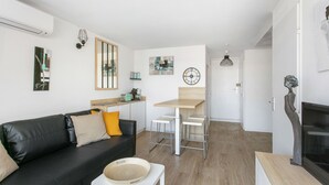 A modern living room with a black leather sofa, a small white table, and a wooden dining table with two stools. There is a fireplace on the right and a kitchen area in the background. - F3 terrace sea view, swimming pool, parking and air conditioning (Sète)