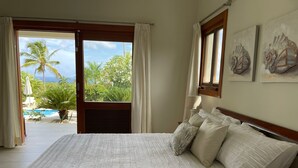 A bedroom with a large bed, a view of a pool and palm trees, and artwork on the wall.