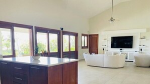 A spacious living area with a large TV, a white sofa, and a kitchen island with a bowl of fruit.