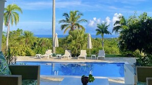 A pool area with lounge chairs and umbrellas, surrounded by palm trees and greenery, with a view of the ocean.