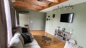 Un salón espacioso con techo alto, vigas de madera, un sofá grande, un televisor y un mueble pequeño.