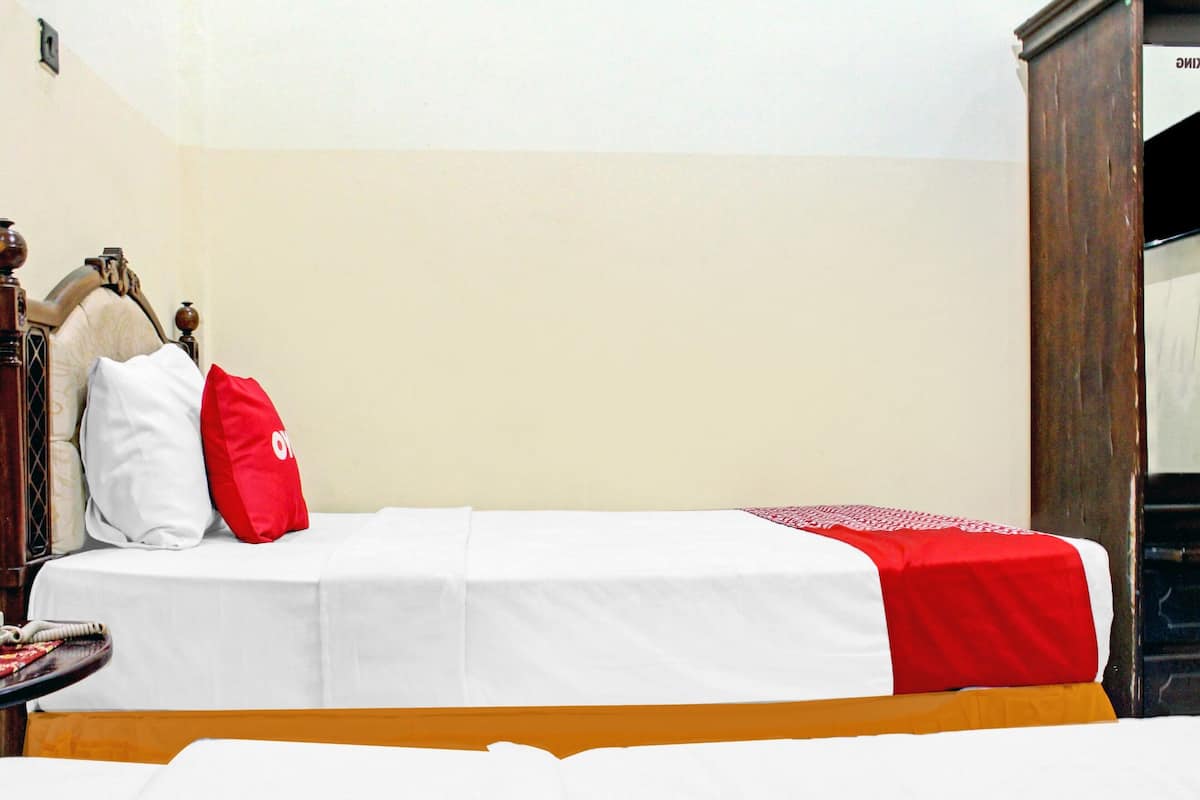 Uma cama de solteiro com colcha branca e vermelha, cabeceira de madeira e uma mesinha de cabeceira com telefone e um objeto vermelho ao lado.