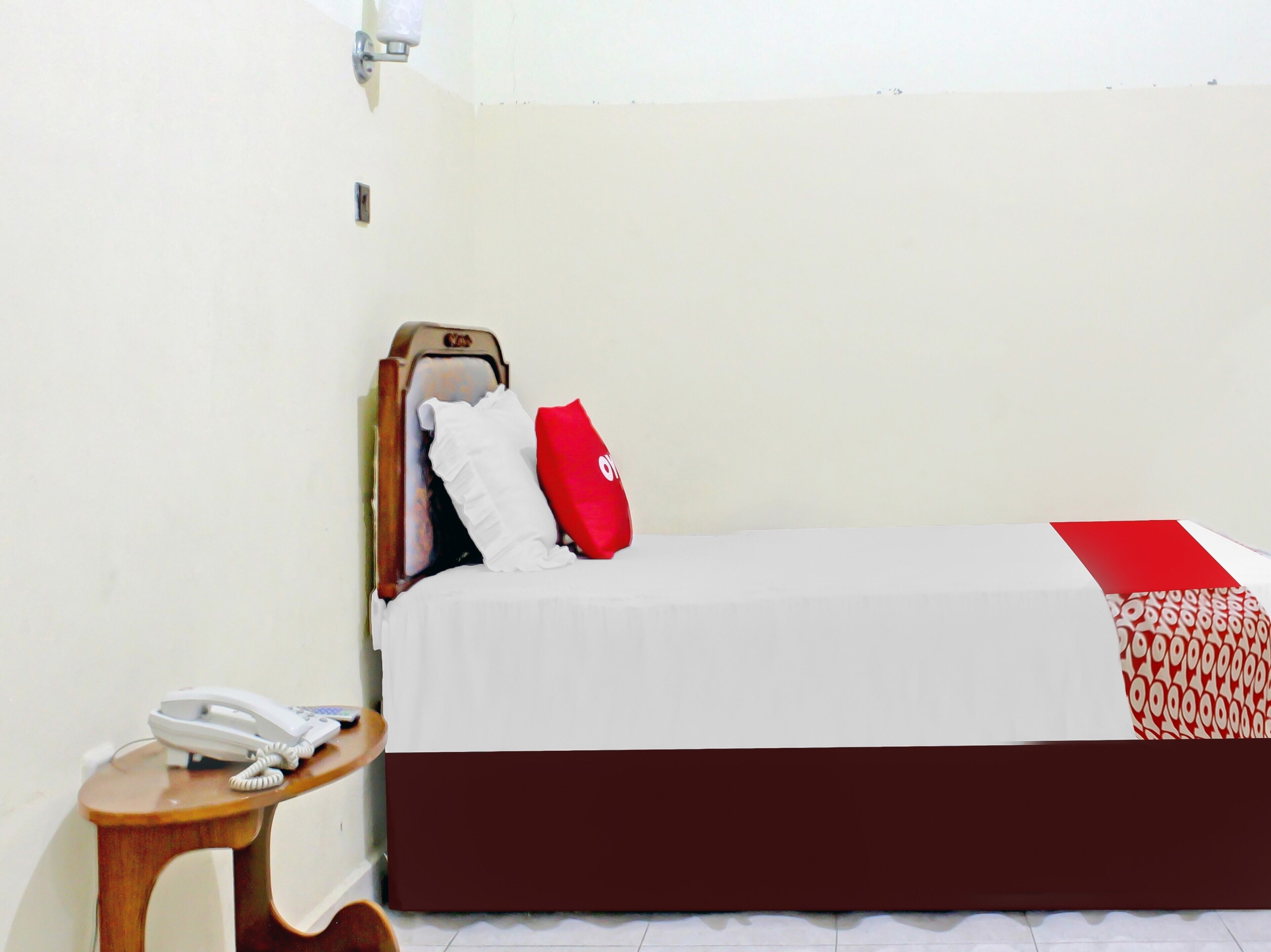 Uma cama de solteiro com um cobreleiro branco e vermelho, um painel de cabeceira em madeira, um travesseiro vermelho e um telefone de mesa branco sobre um criado-mudo de madeira.