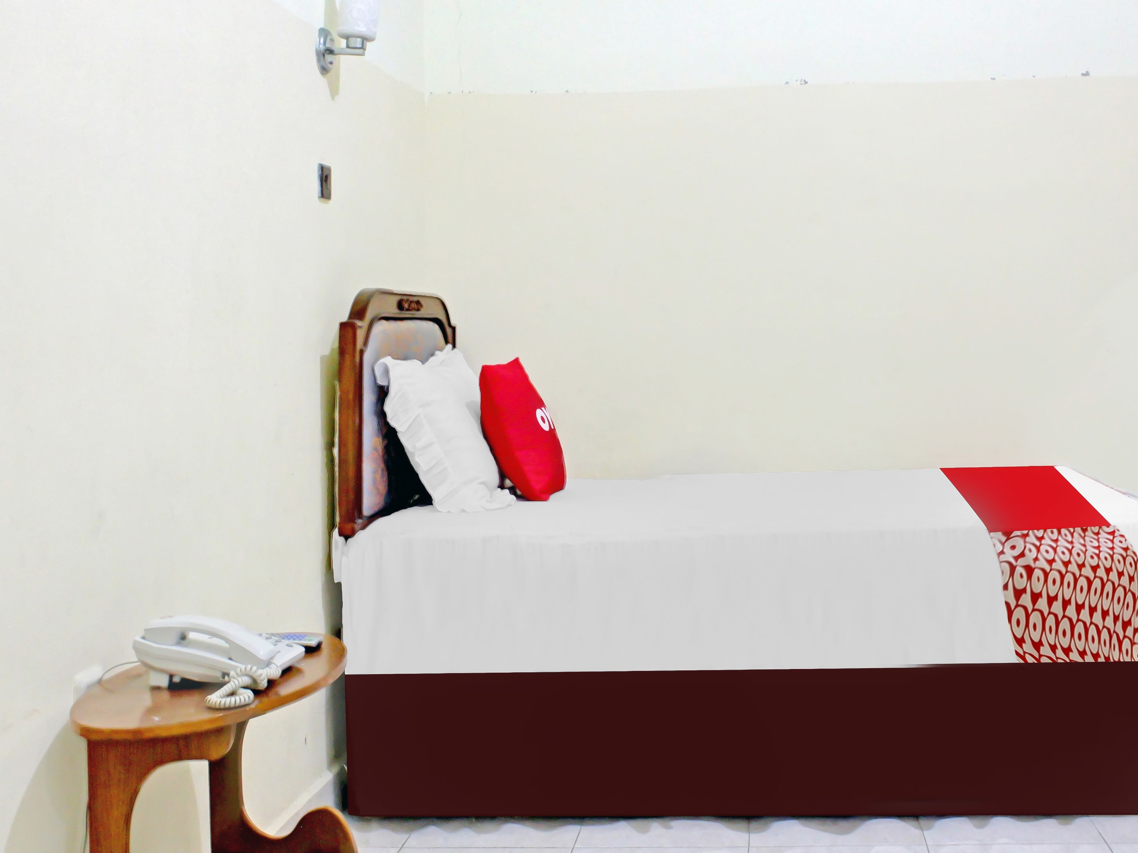A single bed with a white and red bedspread, a wooden headboard, a red pillow, and a white bedside telephone on a wooden nightstand.