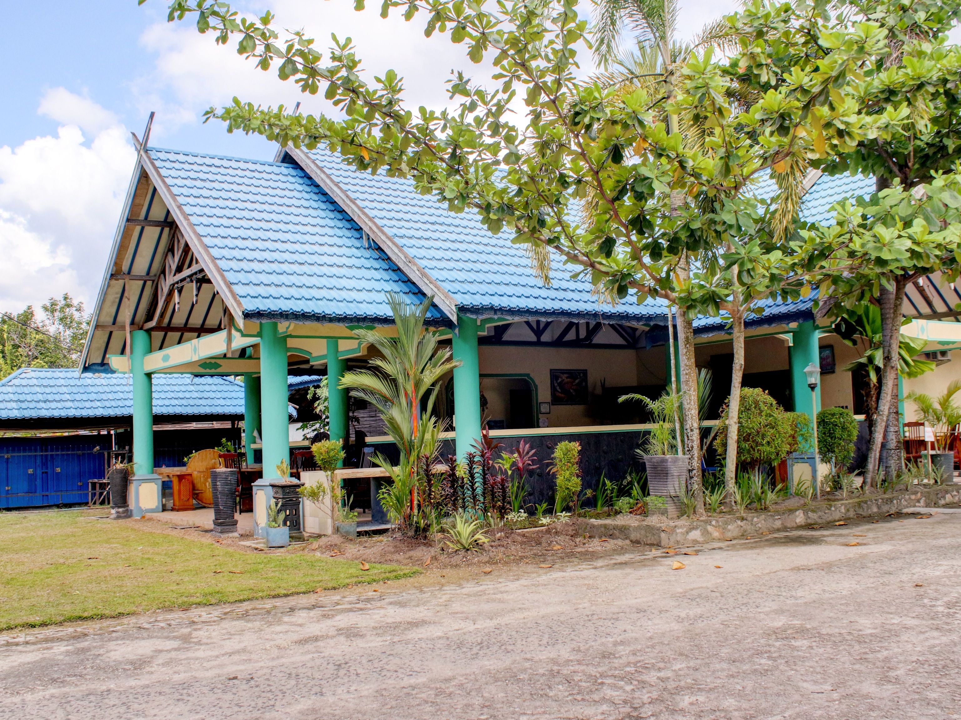 A building with a blue roof and green pillars, surrounded by various plants and a paved area in front.