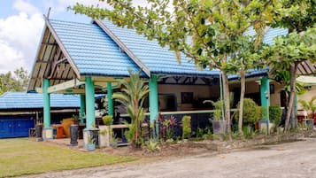 A building with a blue roof and green pillars, surrounded by various plants and a paved area in front.