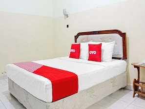 Una cama con una colcha blanca y roja, dos almohadas rojas con el texto 'OYO', un cabecero de madera y una pequeña mesa de madera con un teléfono.