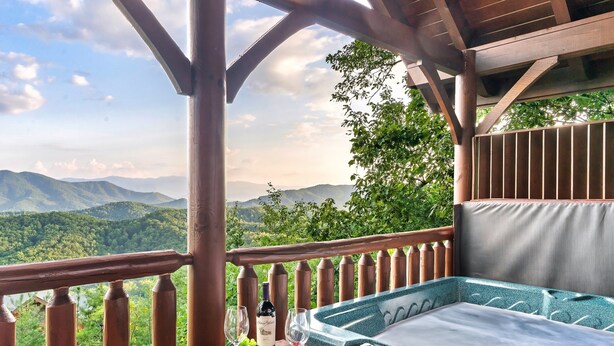 Une terrasse en bois avec un jacuzzi, une assiette de raisins et une bouteille de vin, le tout offrant une vue imprenable sur les montagnes.