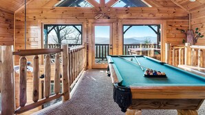 Une cabane en bois dotée d’une table de billard, de rampes en bois et de grandes fenêtres offrant une vue sur les montagnes.