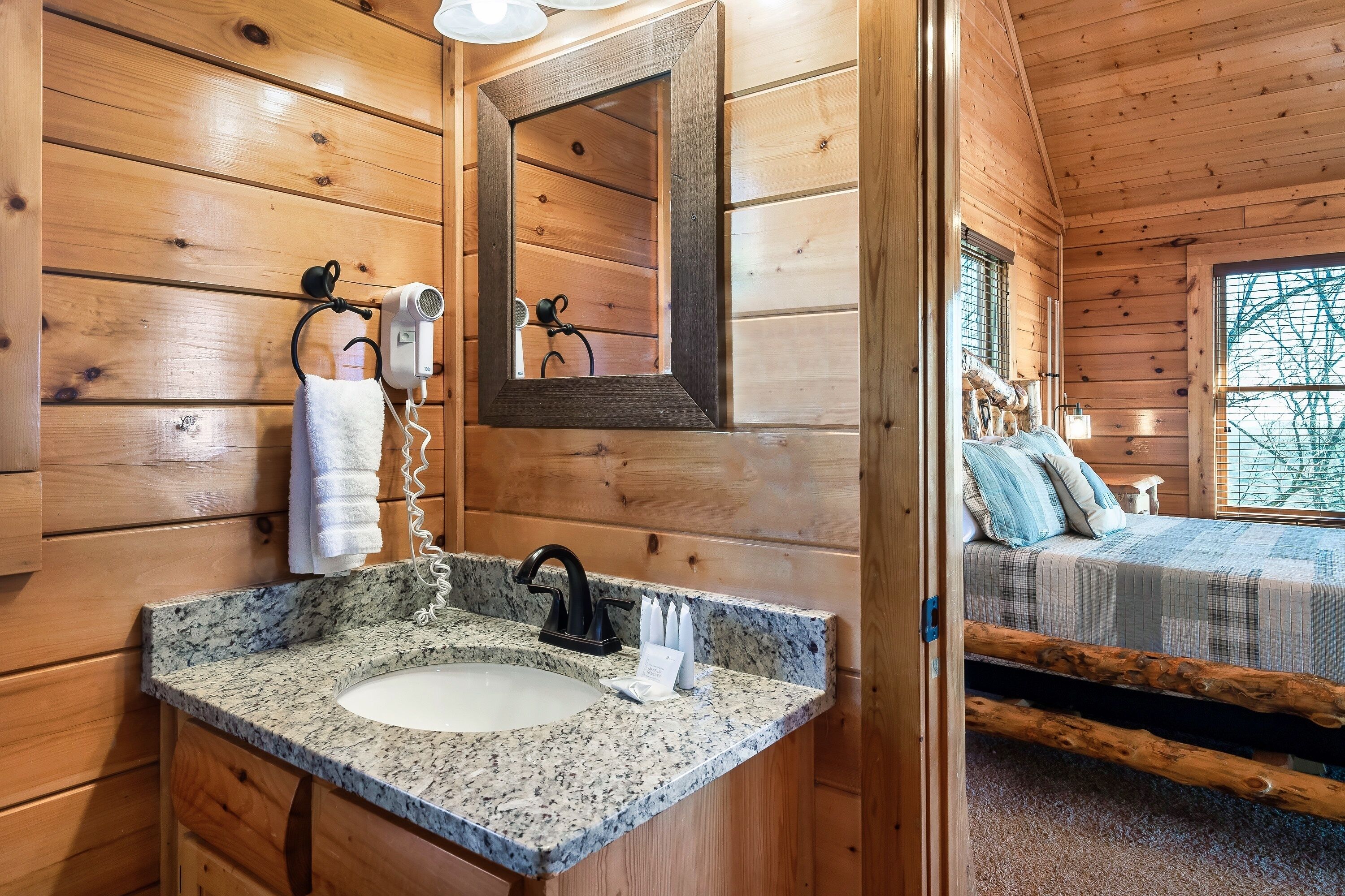 A wooden cabin interior with a granite sink, a mirror, a hairdryer, and a bed with a window view.