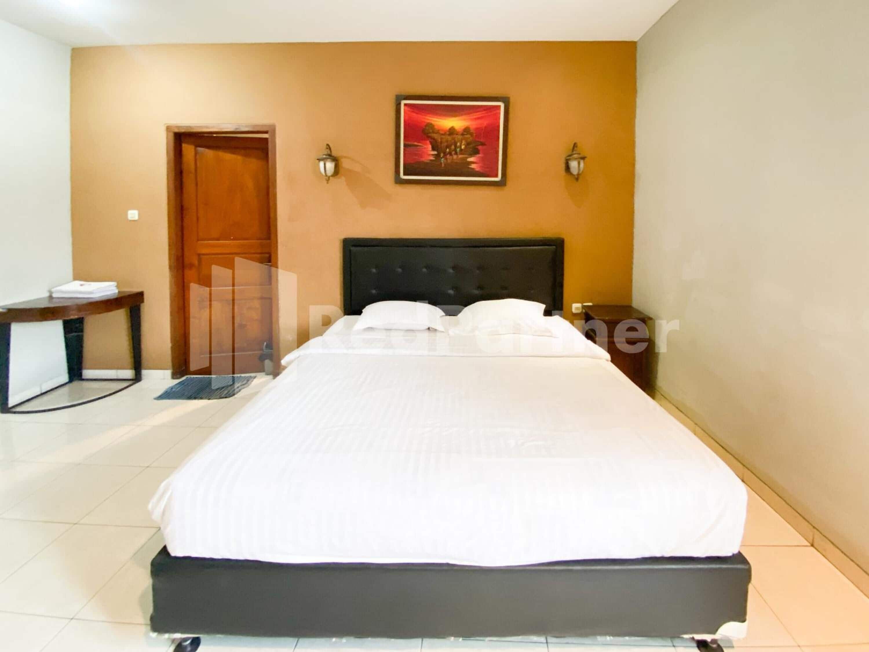 A neatly made bed in a hotel room with a wooden headboard, a desk with a chair, and a door.